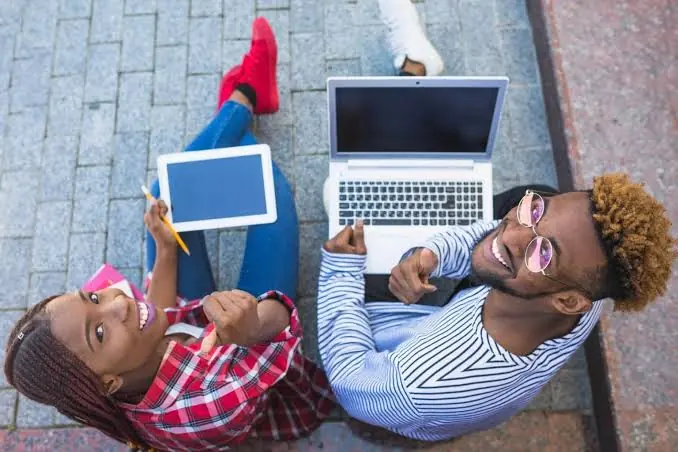 Two students sitting on the pavement with a laptop and a tablet